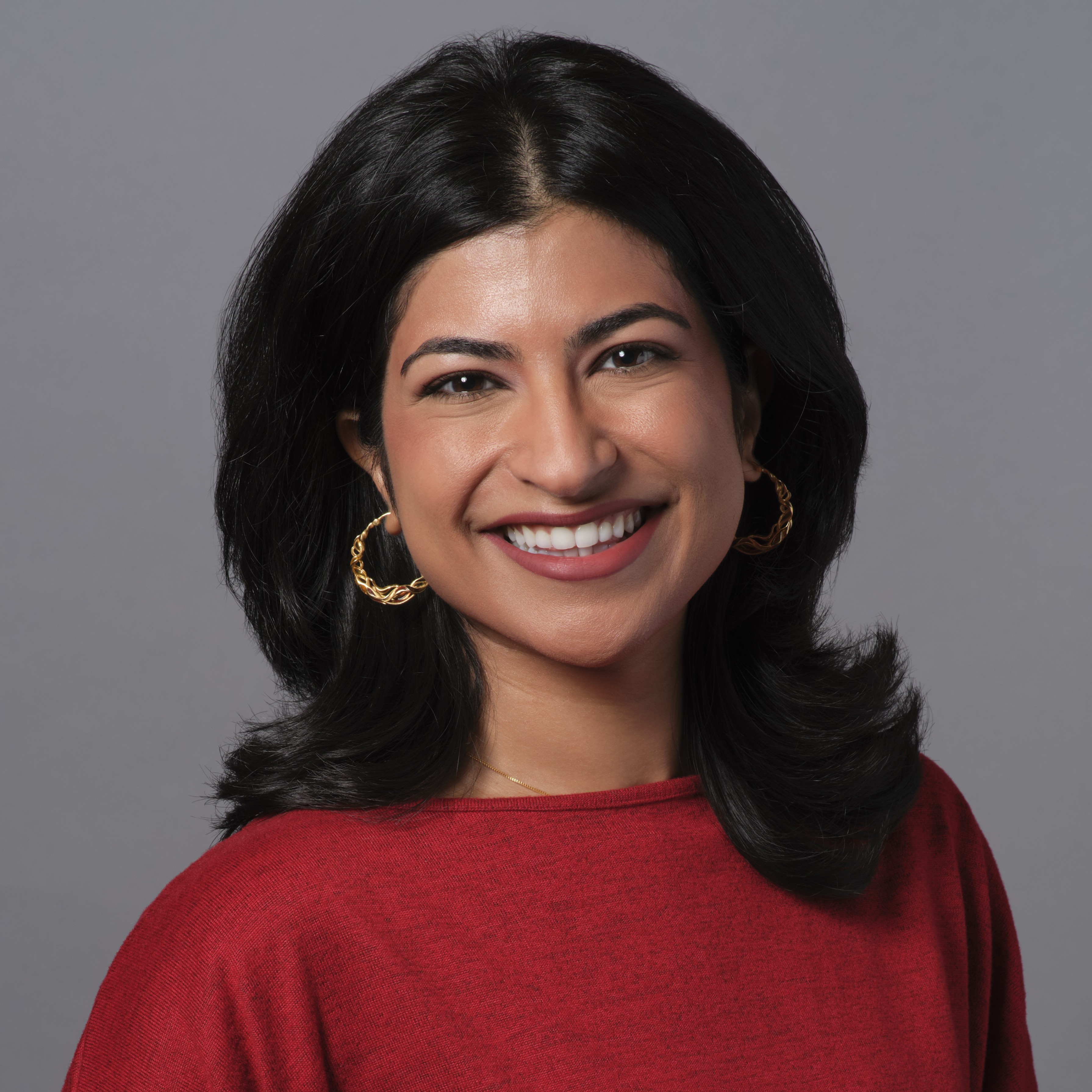 Miti Joshi smiling, wearing a red shirt and gold hoop earrings.